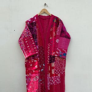 May include: A long, patchwork robe in shades of pink and red, hanging on a wooden hanger. The robe features a variety of patterned fabric squares, stitched together with visible seams. The robe has long sleeves and a relaxed fit.