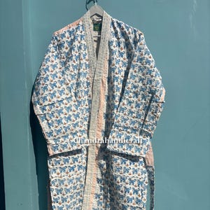 May include: A white robe with a blue animal print and a peach stripe down the center. The robe is hanging on a hanger against a blue background. The robe has a matching belt.