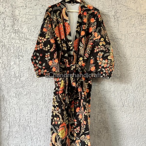 以下が含まれることがあります： オレンジと黄色のアクセントが付いた黒いフローラルプリントのローブ。木製のハンガーに掛けられています。 ローブは、タイクロージャーとストライプの裏地が特徴です。 花柄には葉と花が含まれています。
