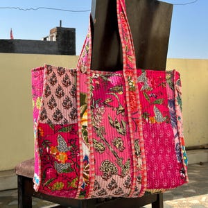 May include: A quilted tote bag with a patchwork design. The bag features various floral and patterned fabrics in shades of pink, red, and white. The bag has sturdy handles and a rectangular shape. The bag is sitting on a wooden chair.