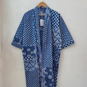 May include: A long, open-front robe with a patchwork design in shades of blue and white. The robe features various geometric and floral patterns, with white trim along the edges. It is hanging on a wooden hanger.