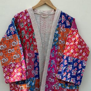 Op de afbeelding: Een patchwork kimono-achtige badjas met een bloemenpatroon in tinten rood, oranje, blauw en roze. De badjas heeft een witte rand en hangt aan een houten hanger.