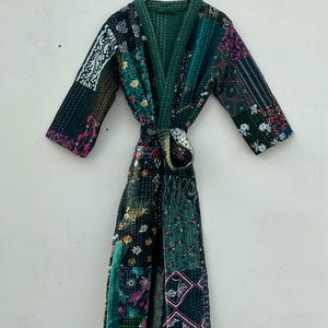 May include: A patchwork robe in shades of green, black, and floral patterns. The robe features a V-neck, long sleeves, and a matching belt. The robe is hanging on a wooden hanger against a white wall.