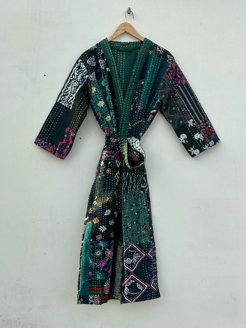 May include: A patchwork robe in shades of green, black, and floral patterns. The robe features a V-neck, long sleeves, and a matching belt. The robe is hanging on a wooden hanger against a white wall.