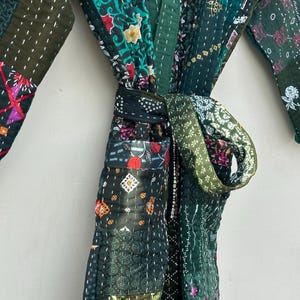 May include: A patchwork robe in shades of green, featuring various floral and geometric patterns. The robe is tied at the waist with a matching fabric belt. The stitching is visible, adding to the handcrafted aesthetic.