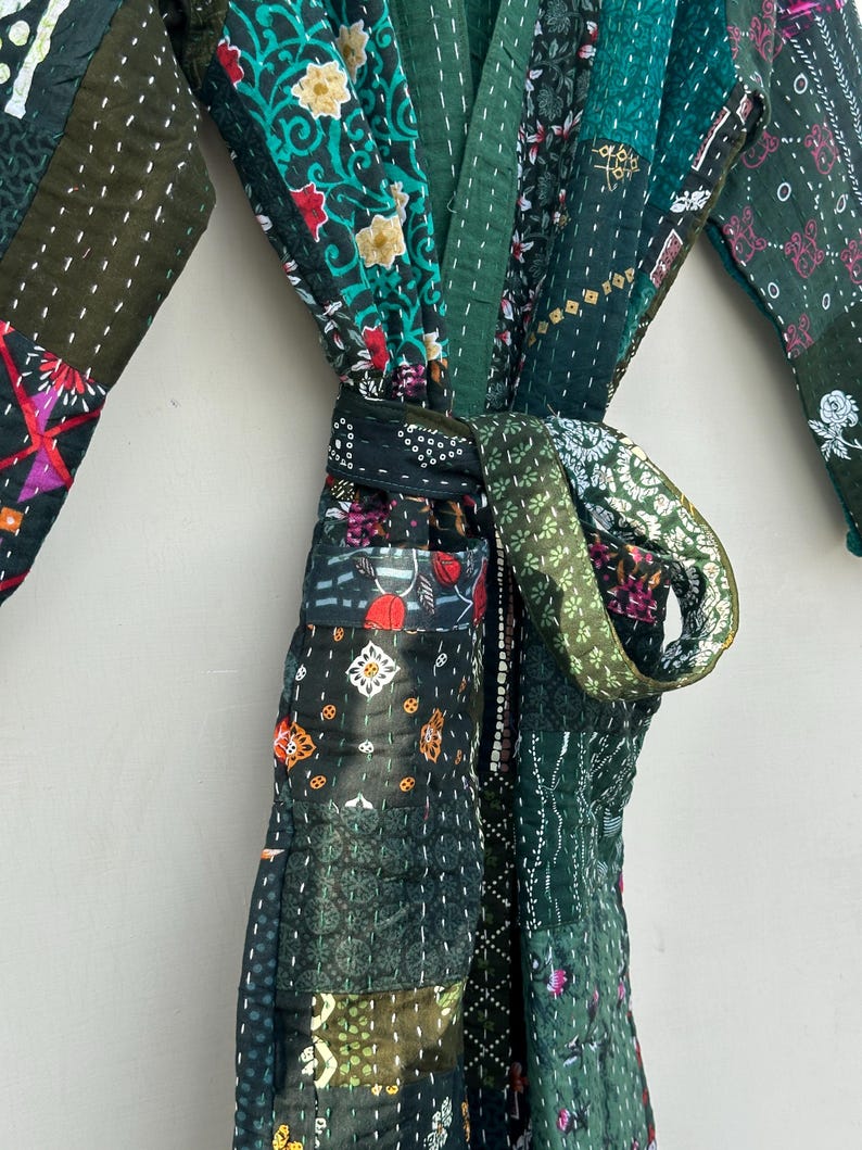 May include: A patchwork robe in shades of green, featuring various floral and geometric patterns. The robe is tied at the waist with a matching fabric belt. The stitching is visible, adding to the handcrafted aesthetic.