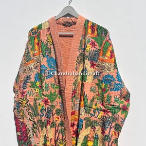 Peut inclure: Une robe matelassée pêche avec un imprimé floral et figuratif vibrant. La robe est suspendue à un cintre gris. L'imprimé comprend du feuillage vert, des fleurs rouges et jaunes et des figures. Le texte "Chandrahandicraft" est visible.