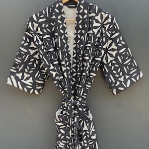 May include: A black and white patterned robe hanging on a wooden hanger. The robe features a repeating white design on a black background. It has a tie closure and pockets. The robe is made of a soft, lightweight fabric.