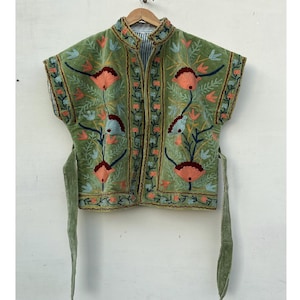 May include: A green embroidered vest with a high collar and short sleeves. The vest features a floral design in shades of orange, blue, and green. It has a tie closure and is hanging on a wooden hanger.