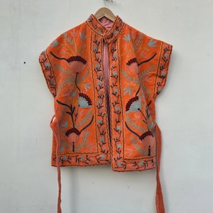 Puede incluir: Chaqueta bordada naranja con diseño floral. La chaqueta tiene mangas cortas y cuello mandarín. El bordado presenta tonos de marrón, azul y verde. La chaqueta tiene cierre de lazo y está colgada de una percha de madera.