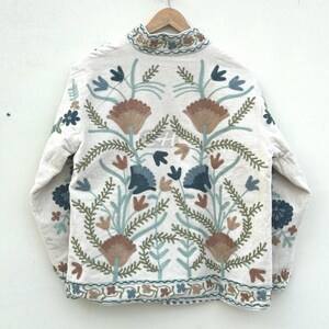 以下が含まれることがあります： クリーム色のジャケット。茶色、青、緑色の複雑な花の刺繍が施されています。ジャケットはハイカラーで、裾と袖口に詳細な刺繍が施されています。デザインには、様式化された花と葉が含まれています。