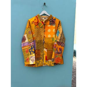May include: A patchwork jacket in shades of yellow and orange with various patterns. The jacket features a mandarin collar, long sleeves, and a button-up front. The fabric is a mix of different textiles, creating a unique and colourful design.