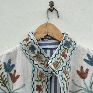以下が含まれることがあります： 青、茶色、緑の色合いの花柄刺繍が施されたクリーム色のジャケット。 ジャケットにはストライプの裏地と木製のハンガーが付いています。 刺繍は襟とフロントエッジを飾っています。