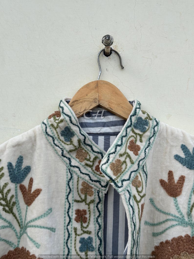 以下が含まれることがあります： 青、茶色、緑の色合いの花柄刺繍が施されたクリーム色のジャケット。 ジャケットにはストライプの裏地と木製のハンガーが付いています。 刺繍は襟とフロントエッジを飾っています。