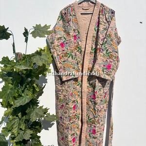 Puede incluir: Bata con estampado floral en tonos rosa, verde y amarillo, colgada de una percha gris. La bata presenta un dise&ntilde;o acolchado con un ribete rosa claro y un cintur&oacute;n a juego. La marca de agua Chandrahandicraft es visible.