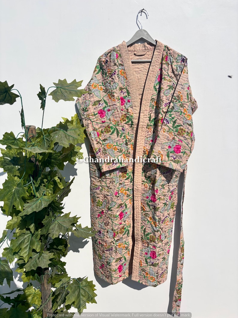 Puede incluir: Bata con estampado floral en tonos rosa, verde y amarillo, colgada de una percha gris. La bata presenta un dise&ntilde;o acolchado con un ribete rosa claro y un cintur&oacute;n a juego. La marca de agua Chandrahandicraft es visible.