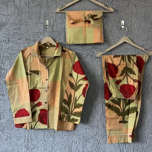 May include: A three-piece pajama set with a floral pattern. The set includes a button-down shirt, pants, and a matching pouch, all hanging on wooden hangers. The fabric features a checkered background with large red flower and green leaf designs.