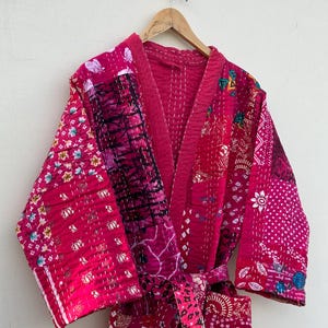 Pode incluir: Um roupão de manga comprida com um design de patchwork em vários tons de rosa e magenta. A peça é feita de tecido acolchoado com detalhes de costura branca visíveis. Inclui um cinto a condizer para atar na cintura.
