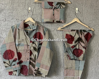 May include: A three-piece pajama set with a floral pattern. The set includes a button-down shirt, pants, and a matching pouch, all hanging on wooden hangers against a textured gray wall. The fabric features red, teal, and brown floral designs.