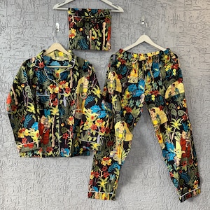 May include: A black pajama set with a vibrant floral and figure print. The set includes a button-down shirt, pants, and a matching eye mask, all hanging on white hangers. The design features colorful flowers, figures, and foliage.