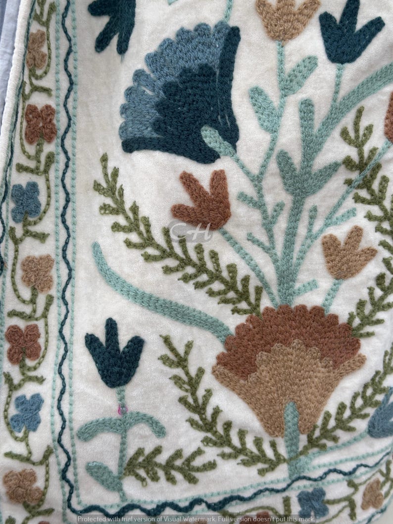 以下が含まれることがあります： 刺繍された花柄の白いテキスタイルのクローズアップ。デザインは、大きな青と茶色の花、緑の葉、青、茶色、緑の色合いの小さな花のモチーフのボーダーが特徴です。刺繍は詳細で質感があります。