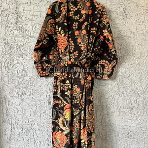 以下が含まれることがあります： オレンジと黄色のアクセントが付いた黒いフローラルプリントのローブ。 ローブは、テクスチャ加工された灰色の壁を背景に、白いハンガーに吊るされています。 ローブは、花と葉の細かいパターンが特徴です。