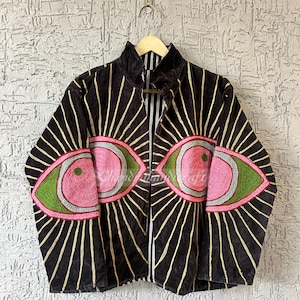 May include: A black velvet zip-up jacket with gold vertical stripes and large embroidered eye designs in pink, green, and blue. The jacket is hanging on a wooden hanger against a textured wall.