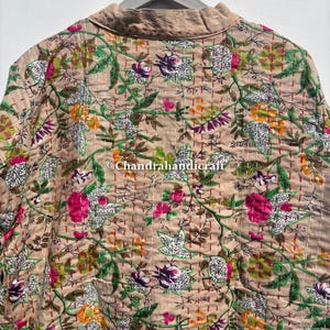 Puede incluir: Chaqueta beige con estampado floral y cuello, colgada de una percha gris. La chaqueta presenta un patr&oacute;n repetido de flores rosas, moradas, naranjas y verdes, con hojas y tallos blancos. Las costuras son rojas.