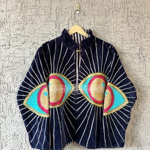 May include: Navy blue jacket with a high collar and a zip-up front. The jacket features two large eye designs with gold, turquoise, and pink embellishments. White vertical lines radiate from the eyes.