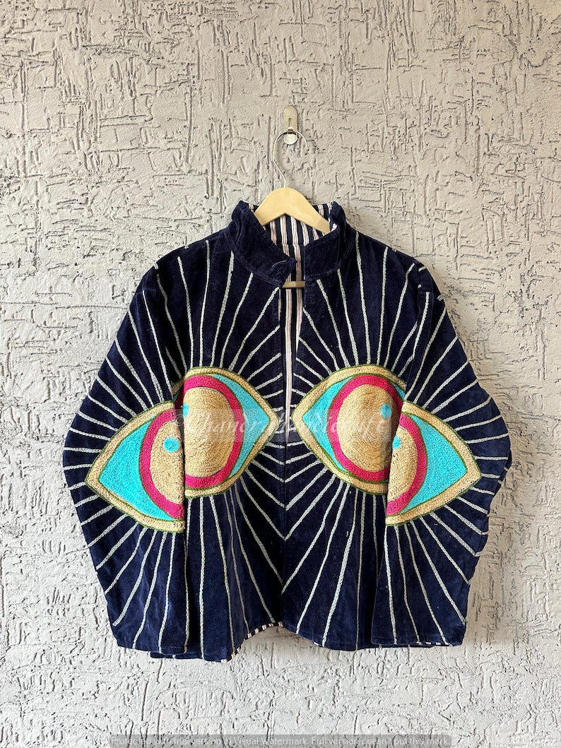 May include: Navy blue jacket with a high collar and a zip-up front. The jacket features two large eye designs with gold, turquoise, and pink embellishments. White vertical lines radiate from the eyes.