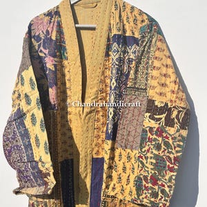 May include: A patchwork kimono-style robe in shades of yellow, blue, and burgundy, hanging on a hanger. The robe features a variety of patterned fabric patches, with visible stitching. The text "Chandrahandicraft" is visible on the robe.