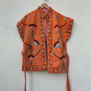 May include: An orange embroidered jacket with a floral design. The jacket has short sleeves and a mandarin collar. The embroidery features shades of brown, blue, and green. The jacket has a tie closure and is hanging on a wooden hanger.