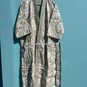Op de afbeelding: Een lange, bedrukte kimono in grijstinten en wit, hangend aan een houten hanger. De kimono heeft een bloemenpatroon en een effen groene rand langs de randen en de voorkant. De tekst "Chandrahandicraft" is zichtbaar.