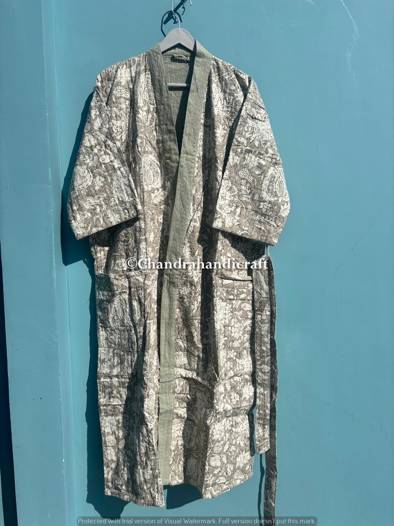 Op de afbeelding: Een lange, bedrukte kimono in grijstinten en wit, hangend aan een houten hanger. De kimono heeft een bloemenpatroon en een effen groene rand langs de randen en de voorkant. De tekst "Chandrahandicraft" is zichtbaar.