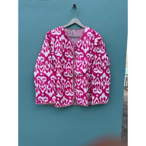 May include: A pink and white patterned jacket with a V-neck and long sleeves, suspended from a wooden hanger. The jacket features a reversible design with a striped interior and decorative trim.