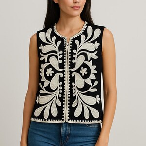 May include: Black sleeveless vest featuring white floral embroidery. The vest has a zip-up front and a round neckline. The white embroidery provides a strong contrast against the black fabric. The vest is worn with blue jeans.