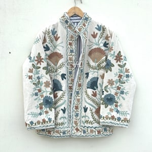 Op de afbeelding: Witte jas met ingewikkelde bloemenborduurwerk in blauw, bruin en groen. De jas heeft een opstaande kraag en is versierd met gedetailleerde stiksels langs de randen. De jas hangt aan een houten hanger.