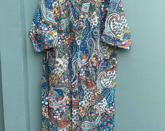 May include: A long, open-front robe with a vibrant paisley pattern in shades of blue, orange, yellow, and white. The robe is hanging on a wooden hanger against a teal wall. The robe has short sleeves and a tie.