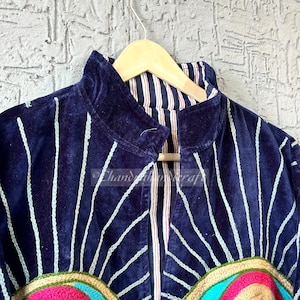 May include: A navy blue velvet jacket with white vertical lines and colorful embroidered details. The jacket has a high collar and is hanging on a wooden hanger. The embroidery includes pink, blue, and tan colors.