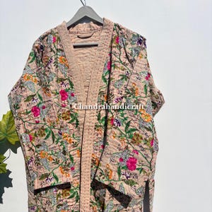 May include: A floral-patterned robe in shades of beige, pink, green, and orange hangs on a gray hanger. The robe features a quilted design with a floral print. The robe is made of a soft, lightweight fabric.