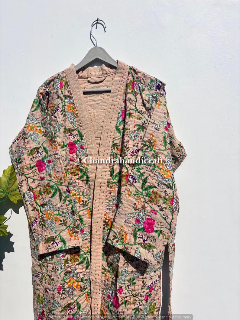 Puede incluir: Una bata con estampado floral en tonos beige, rosa, verde y naranja cuelga de una percha gris. La bata presenta un dise&ntilde;o acolchado con un estampado floral. La bata est&aacute; hecha de una tela suave y ligera.