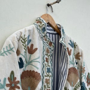 以下が含まれることがあります： 茶色、ティール、青の色合いの花柄が刺繍されたクリーム色のジャケット。 ジャケットは、明るい色の壁に木製のハンガーに掛けられています。 内側の裏地は青と白のストライプです。