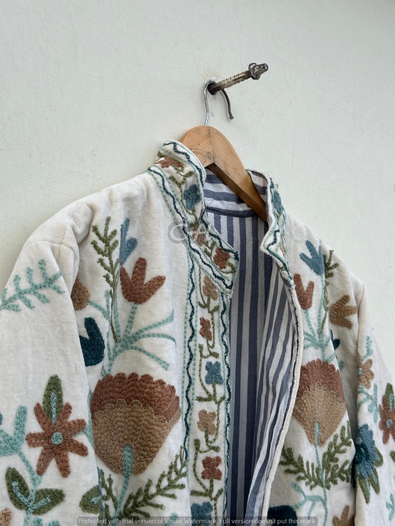 以下が含まれることがあります： 茶色、ティール、青の色合いの花柄が刺繍されたクリーム色のジャケット。 ジャケットは、明るい色の壁に木製のハンガーに掛けられています。 内側の裏地は青と白のストライプです。