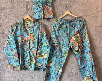 Puede incluir: Un conjunto de pijama azul claro con un estampado floral, que incluye una camisa con botones, pantalones y una bolsa a juego. El diseño floral presenta acentos rojos, verdes y amarillos. El conjunto se exhibe en perchas de madera contra una pared gris texturizada.