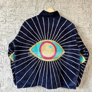 May include: A navy blue jacket with a large, embroidered eye design on the back. The eye features turquoise, pink, and gold accents, with silver rays emanating outwards. The jacket is hanging on a wooden hanger.