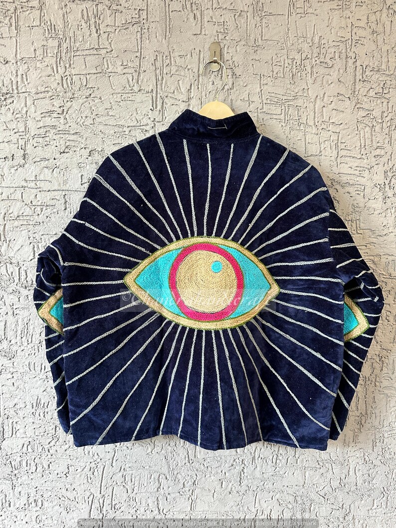 May include: A navy blue jacket with a large, embroidered eye design on the back. The eye features turquoise, pink, and gold accents, with silver rays emanating outwards. The jacket is hanging on a wooden hanger.