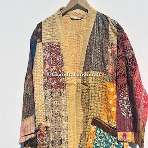 Pode incluir: Um roupão estilo quimono em patchwork com gola e painéis frontais bege. O roupão é feito de vários retalhos de tecido em tons de bege, marrom, verde-azulado e bordô. O roupão está pendurado em um cabide de madeira em um fundo branco.