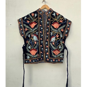 Puede incluir: Chaqueta sin mangas bordada en azul marino con cuello mandarín. La chaqueta presenta un diseño floral en tonos coral, azul y dorado. Tiene cierres de lazo y está colgada de una percha de madera.