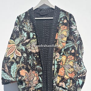 Könnte beinhalten: Schwarze Kimono-Jacke mit floralem Druck und Steppmuster. Die Jacke hat ein lebendiges Muster aus Blumen und Blättern in Orange-, Grün- und Gelbtönen. Die Innenseite ist einfarbig schwarz mit weißen Nähten. Die Jacke hängt an einem Holzkleiderbügel.