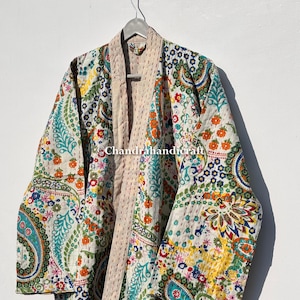 以下が含まれることがあります： ペイズリーと花柄のデザインが施された、模様入りの着物ローブ。ローブは、青、オレンジ、緑、黄色の鮮やかなアクセントが施されたクリーム色のベースが特徴です。ローブは白い背景に金属製のハンガーに吊るされています。